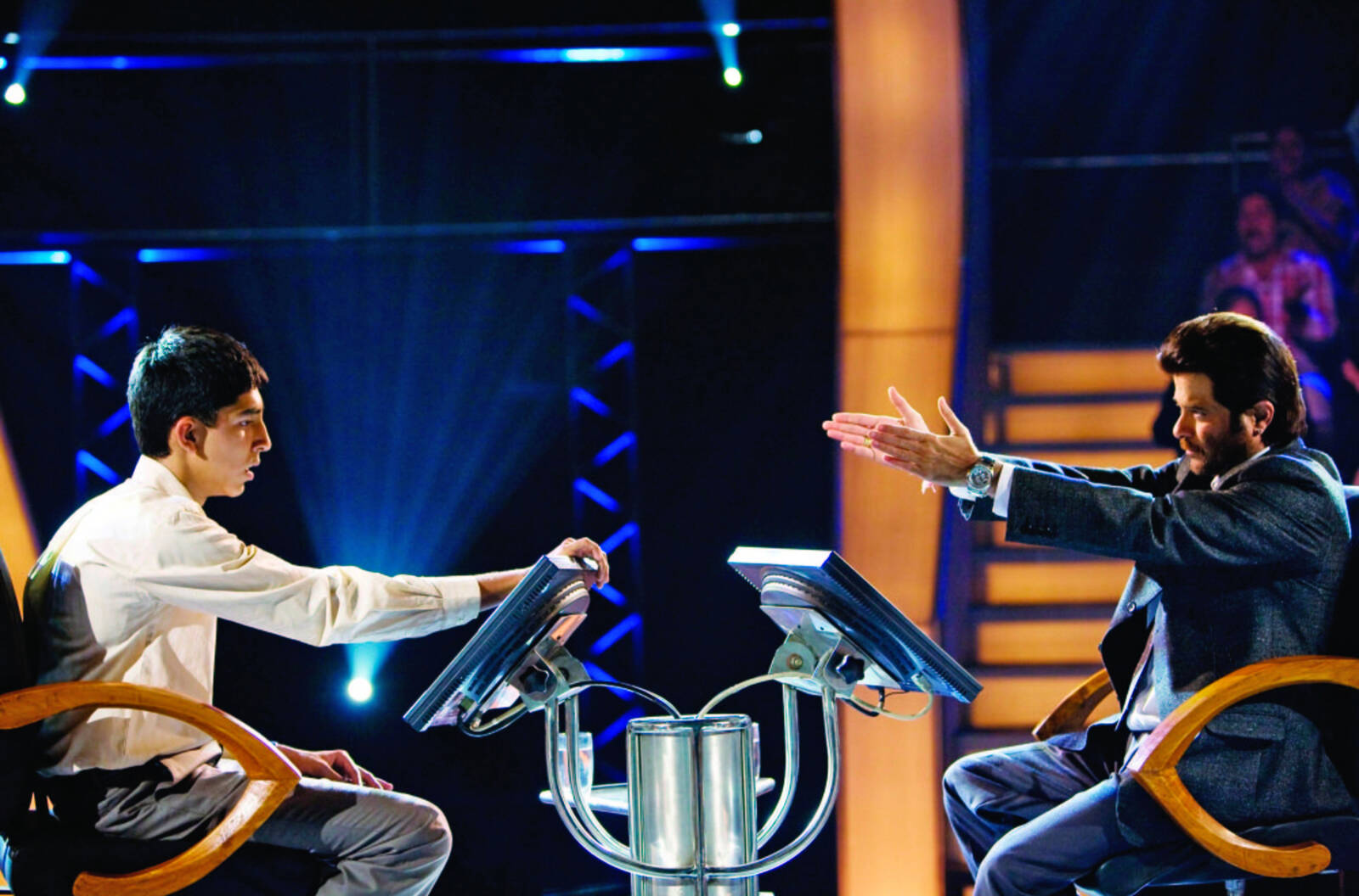 Schauspieler Dev Patel als Jamal und Anil Kapoor als Moderator sitzen auf den Quizstühlen bei „Wer wird Millionär?“