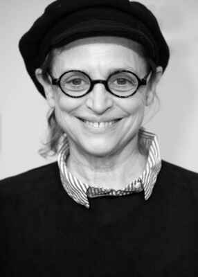 Katharina Thalbach lächelt mit Brille und Hut in einem schwarz/weiß Foto.