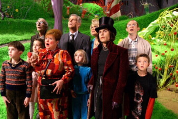 Johnny Depp als Willy Wonka mit bleicher Haut und Zylinder, zusammen mit der Gruppe an Kindern und Begleitpersonen auf einer buntern Wiede in der Schokoladenfabrik.