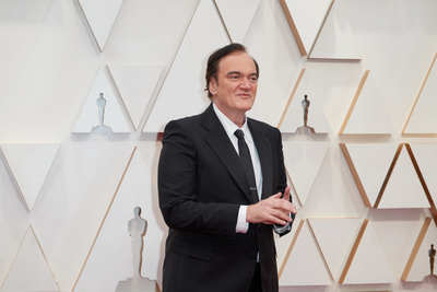 Quentin Tarantino „Cinema Speculation“: Zu sehen ist Tarantino in einem schwarzen Anzug, mit weißem Hemd und schwarzer Krawatte vor der Oscar Wand (2020).