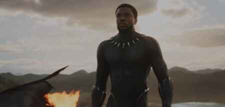 Man sieht Chadwock Boseman im Kostüm vim Black Panther, einen hautengen schwarzen Anzug; im Hintergrund liegt eine brennende Flugkapsel, der Himmel färbt sich langsam orange.