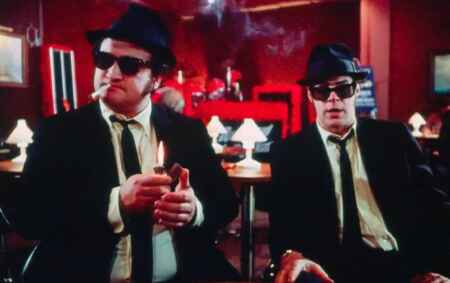 Dan Aykroyd (l.) und John Belushi in „Blues Brothers“: Beide tragen ein weißes Hemd, mit schwarzer Krawatte, Anzug und Hut, sowie eine Sonnenbrille, sie sitzen am Tisch in einer rot beleuchteten Bar.