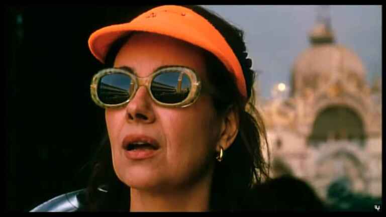 Licia Maglietta als Rosalba in Nahaufnahme mit Sonnenbrille und orangenem Cappie, sie schaut beeindruckt Venedig an, im Hintergrund ist ein antiker Kuppelbau zu sehen.