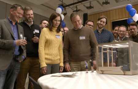 „Downsizing“: Man sieht eine Gruppe an normalgroßen Menschen, im Vordergrund Paul (Matt Damon) unf Audrey (Kristen Wiig), die an einem Tisch stehen, wo zwei Miniatur-Menschen mit Megafon drauf stehen.