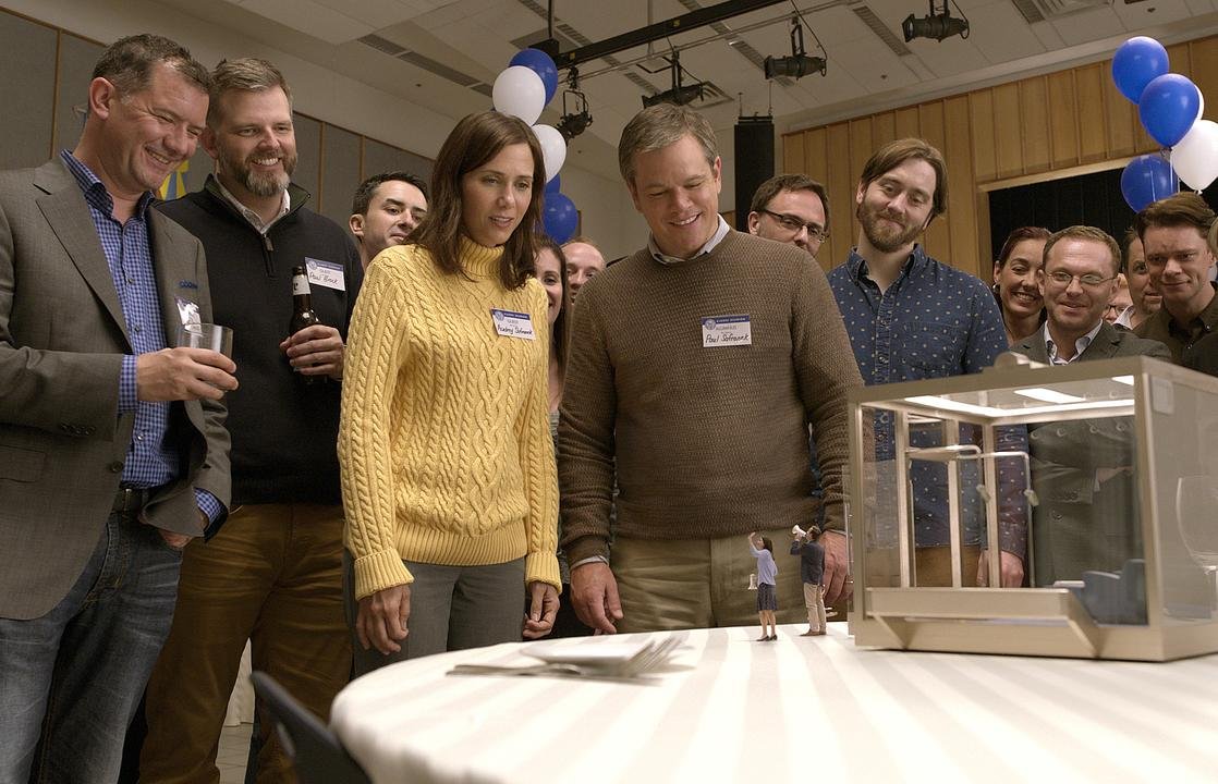 „Downsizing“: Man sieht eine Gruppe an normalgroßen Menschen, im Vordergrund Paul (Matt Damon) unf Audrey (Kristen Wiig), die an einem Tisch stehen, wo zwei Miniatur-Menschen mit Megafon drauf stehen.