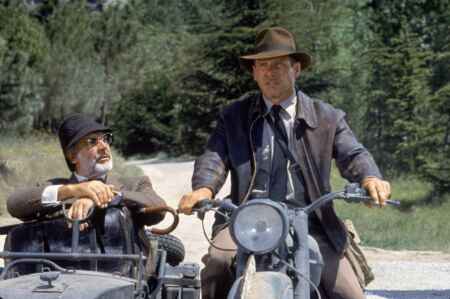 Harrison Ford als Indiana Jones mit Hemd, Krawatte, Lederjacke und Hut auf einem Motorad, Sean Connery (l.) alias Prof. Henry Jones mit Hemd, Krawatte und Jacket sitzt im Beiwagen.