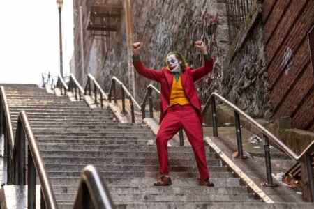 „Joker 2“: Man sieht Joaquin Phoenix als Joker, in einem roten Anzug, mit zurückgegelten Haaren; sein Gesicht ist weiß geschminkt, mit einem großen roten, lachenden Mund, einer roten Nase und Rauten über den Augen; er tanzt mit nach oben gehobenen Armen auf einer öffentlichen Treppe.