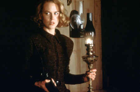 „The Others“: Man sieht Nicole Kidman in einem hochgeschlossenen schwarzen Kleid mit einer alten Öllampe und einer Waffe in den Händen, sie schaut verängstigt und aufmerksam.