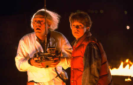 „Zurück in die Zukunft“: Man sieht Dr. Brown (Christopher Lloyd, l.) und Marty (Michael J. Fox) geblendet von hellem Licht und Doc hält eine Art Fernbedienung in der Hand, im Hintergrund ist ein kleines Feuer.