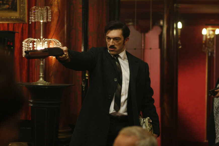 Vincent Cassel als Jacques Mesrine mit einer Waffe in deiner ausgestreckten Han und im Anzug in einem noblen Restaurant.