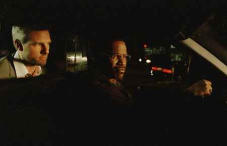 Tom Cruise (l.) sitzt im hellen Anzug auf der Rückbank eines Taxis, in dem er dunkel ist, Jamie Foxx ist der Taxifahrer und sitzt hinter dem Lenkrad. Beide sind von der (Beifahrer-)Seite aus zu sehen und es ist dunkel im Auto.