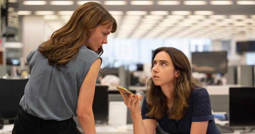 „She Said“-Trailer: Carey Mulligan als Megan Twohey (l.) sitzt auf dem Schreibtisch und Zoe Kazan als Jodi Kantor sitzt am Schreibtisch, mit einem Handy in der Hand auf Lautsprecher gestellt, die beiden schauen sich an, im Hintergrund ist ein Redaktionsbüro zu sehen.