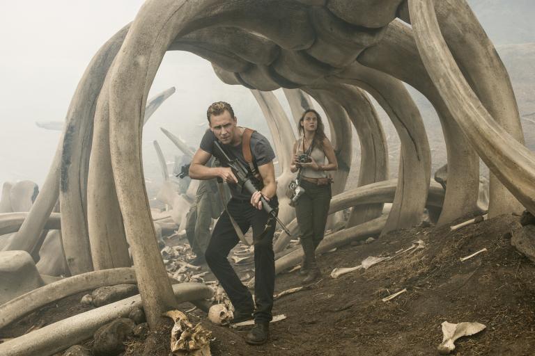 „Kong: Skull Island“ im TV: Man sieht Tom Hiddleston und Brie Larson, wie sie durch ein riesiges Skelett laufen, sie haben Waffen dabei, der Hintergrund ist vernebelt.