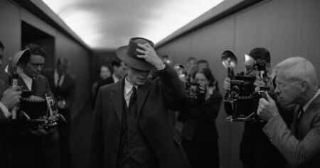 „Oppenheimer“ Teaser: Man sieht Schauspieler Cillian Murphy aöls J. Robert Oppenheimer, er geht durch einen Gang mit Pressemenschen und hat einen Hut in sein Geschichte gezogen; die Aufnahme ist in Schwarz/weiß.