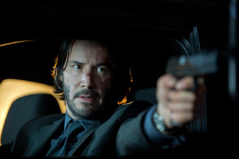 „John Wick“ im TV: Man sieht Keanu Reeves als John Wick in einer Nachaufnahme, wie er im Auto sitzt und durch das offene Fenster mit einer Pistole auf etwas/jemanden hinter der Kamera ziehlt, er trägt den typischen schwarzen Anzug.