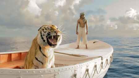 „Life of Pi: Schiffbruch mit Tiger“ im TV: Man sieht ein Rettungsboot auf einem ruhigen Ozean, im voredern Teil sitzt ein begalischer Tiger und fletscht die Zähne, im hinteren Teil steht ein Junge mit geballten Fäusten und ernster Miene.