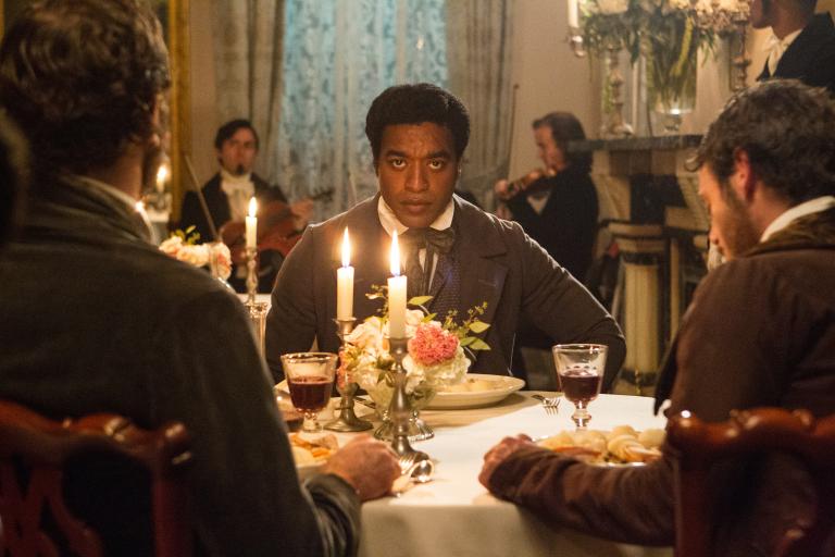 „12 Years a Slave“ im TV: Man sieht rechts und Links 2 Männer mit dem Rücken zur Kamera sitzen, ihnen mittig gegenüber sitzt ein Afroamerikaner in guter Kelidung und schaut zwischen den Männern in die Kamera, sie sind in einem guten Restaurant.