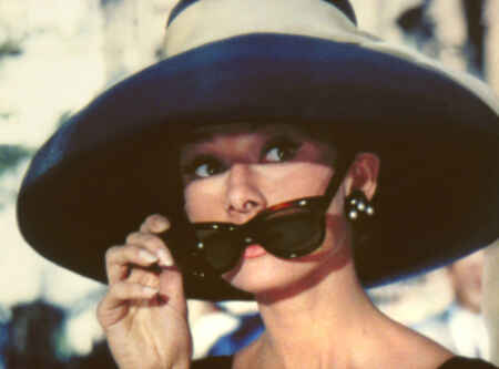 „Frühstück bei Tiffany“ im TV: Man sieht Audrey Hepburn als Holly Golightly in Nahaufnahme, sie trägt einen großen Hut, teure Ohrringe und zieht dieSonnenbrille von ihren Augen weg.