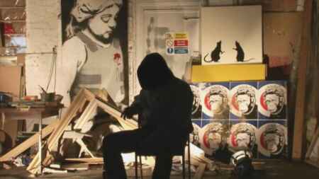 „Banksy – Exit Through the Gift Shop“ im TV: Man sieht eine dunkel gekleidete Person auf einem Stuhl sitzend, im Hintergrund sieht man ein Banksy-Bild, Skizzen und Farben.